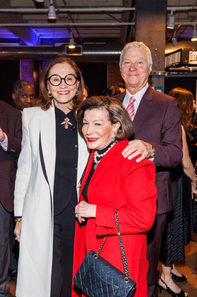 Two women, one in a long white jacket and the other in a long red jacket with a man in a purple suit.