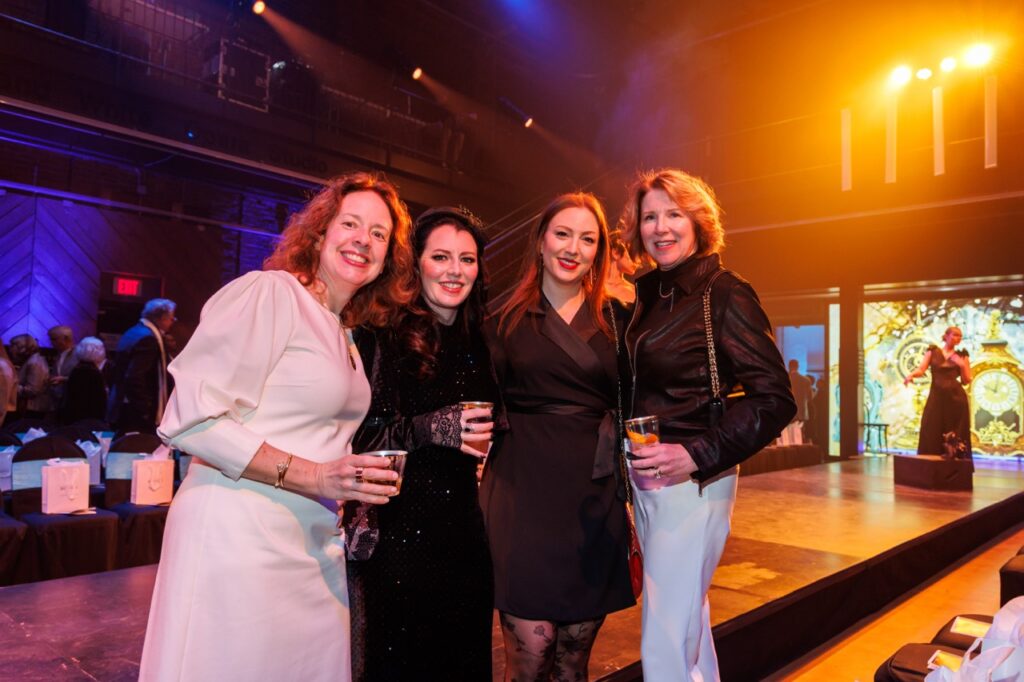 Four women stand with drinks in front of the Pittsburgh Opera catwalk.