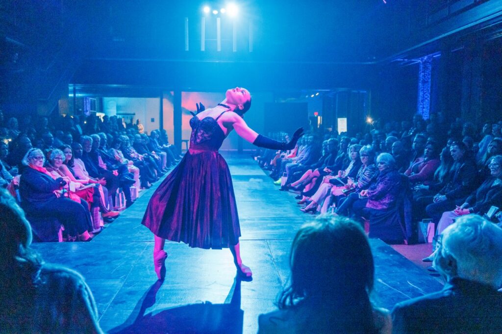 A woman on a catwalk in a lacy black dress, leaning back in the light.