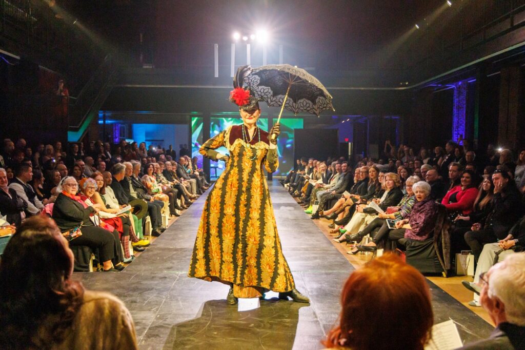 A woman in a yellow flower and black stripe dress with a fancy fascinator hat and umbrella.