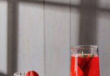 Juneteenth Red Punch (Berry Peach Lemonade) Three glasses of red punch beside a large pitcher of the punch with mint leaves inside and strawberries on the rim.