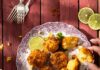 Mexican Street Cornbread A person picks up a Mexican Street Cornbread fritter off a plate that sits beside slices limes, three forks, an ear of corn, and a bowl of lime crema with cilantro.