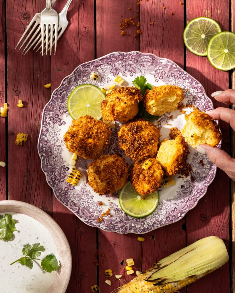 A person picks up a Mexican Street Cornbread fritter off a plate that sits beside slices limes, three forks, an ear of corn, and a bowl of lime crema with cilantro.