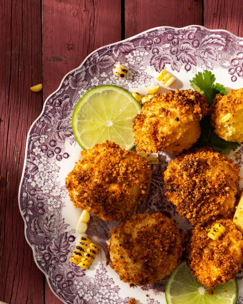 A plate with a burgundy pattern around the edge with little Mexican Street Cornbread fritters and slices of limes.