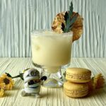 A short glass with a creamy yellow cocktail and a dehydrated pineapple slice behind a Minions mummy figure and two yellow macarons.