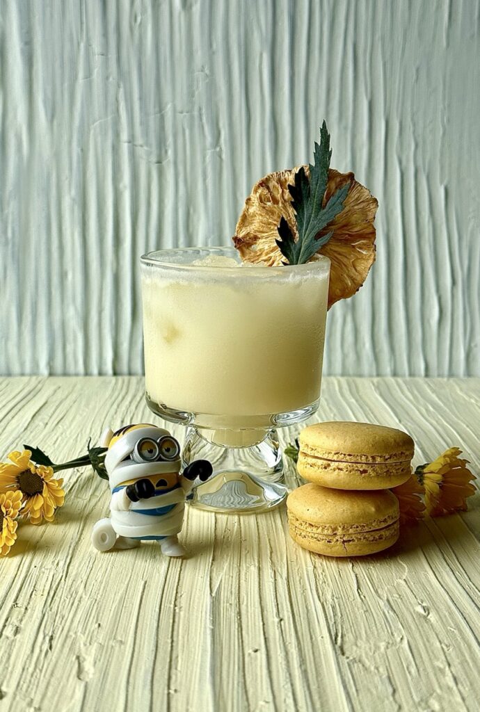 A short glass with a creamy yellow cocktail and a dehydrated pineapple slice behind a Minions mummy figure and two yellow macarons.
