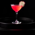 A Nick and Nora glass with a bright magenta dragon fruit cocktail inside with a dehydrated dragon fruit slice garnish.