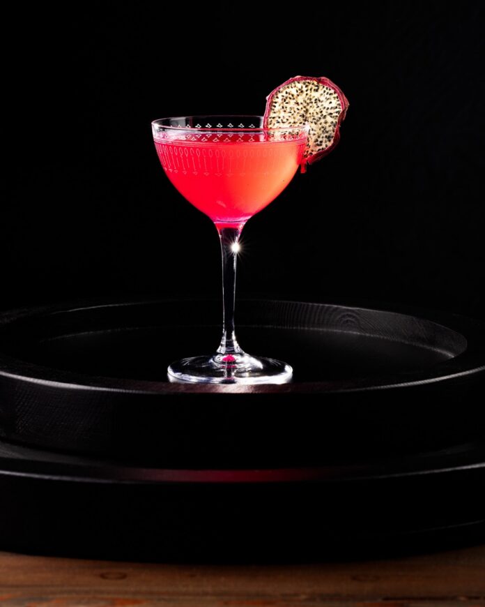 A Nick and Nora glass with a bright magenta dragon fruit cocktail inside with a dehydrated dragon fruit slice garnish.