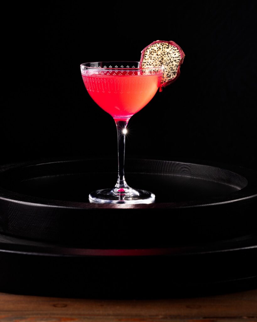 A Nick and Nora glass with a bright magenta dragon fruit cocktail inside with a dehydrated dragon fruit slice garnish.