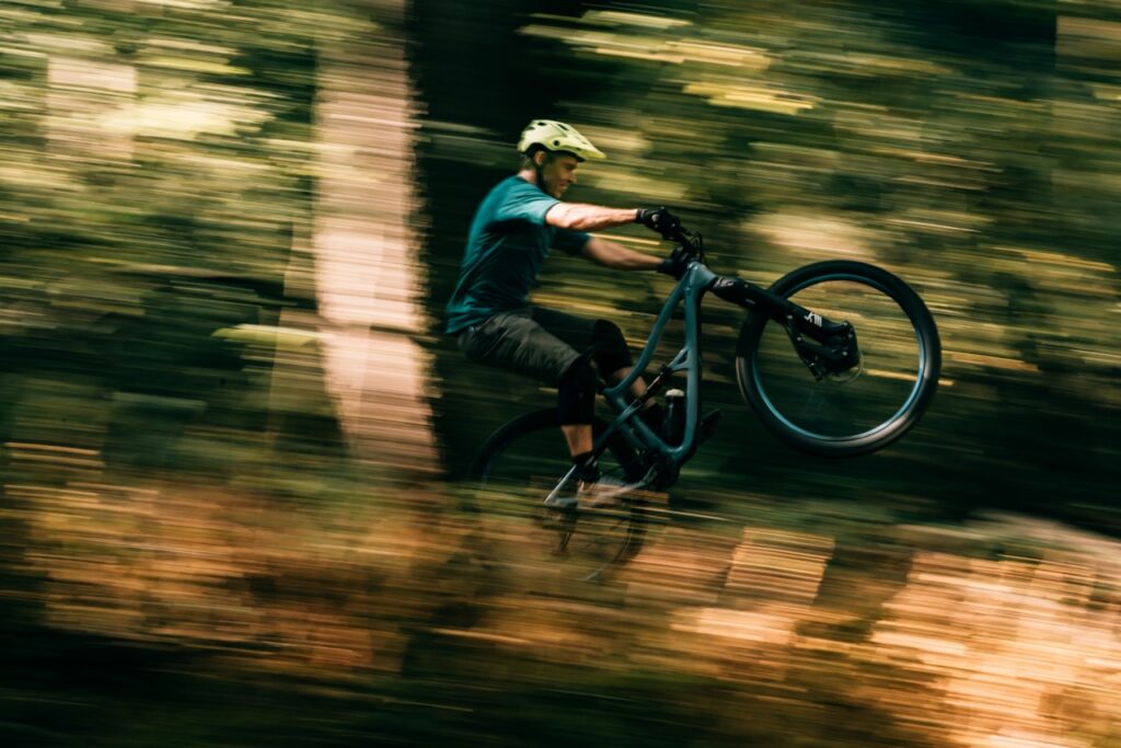 A man mountain bikes through the forest with his front wheel popped up.