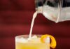 Craving a New Summer Cocktail? Here’s What Pittsburgh’s Bartenders Recommend A yellow cocktail being poured from a shaker into a rocks glass with an orange peel garnish all on a red background.