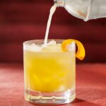 A yellow cocktail being poured from a shaker into a rocks glass with an orange peel garnish all on a red background.