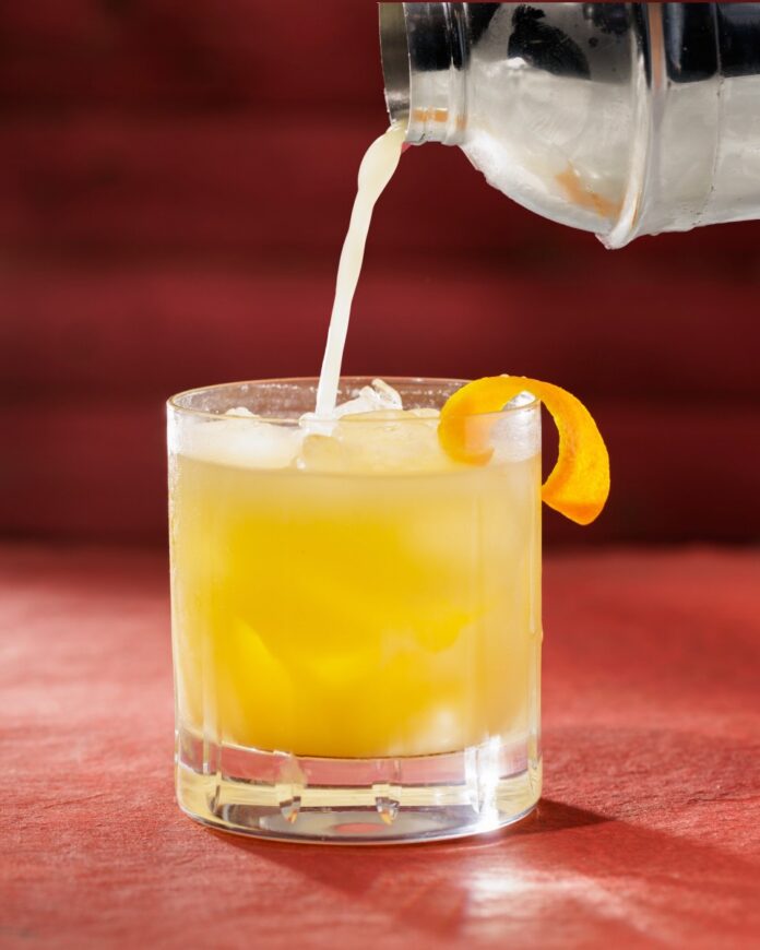 A yellow cocktail being poured from a shaker into a rocks glass with an orange peel garnish all on a red background.