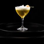 A lychee cocktail with a foam on top in a martini glass on a black tray.