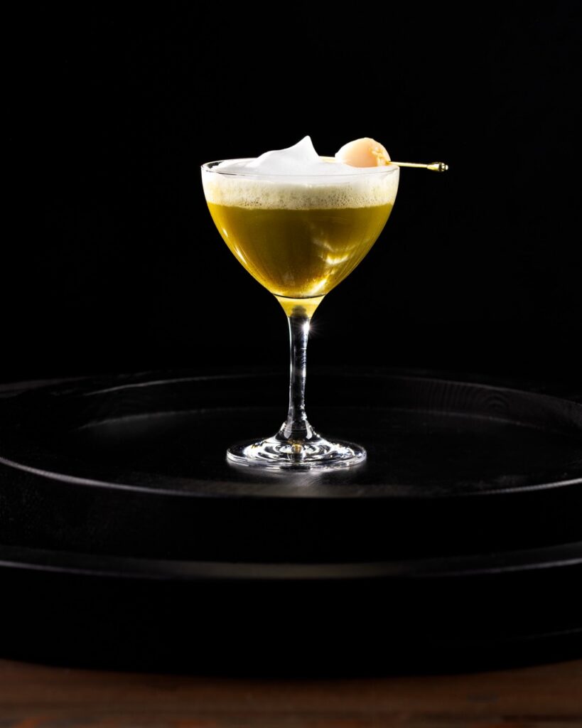 A lychee cocktail with a foam on top in a martini glass on a black tray.