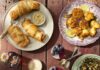 What’s Delicious for Summer? Picnic Recipes from Chef Kenny Cumberland A picnic table holds five plates of food with pigs in a blanket, chicken sandwiches, street cornbread, grain bowl, sauerkraut, two cups of cakes and a glass of tea punch.