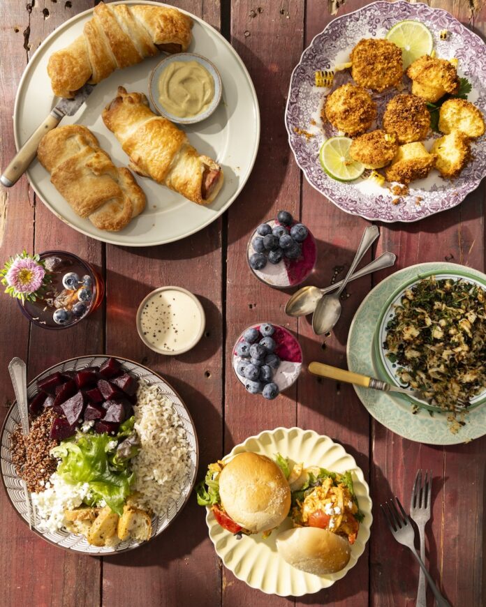 A picnic table holds five plates of food with pigs in a blanket, chicken sandwiches, street cornbread, grain bowl, sauerkraut, two cups of cakes and a glass of tea punch.