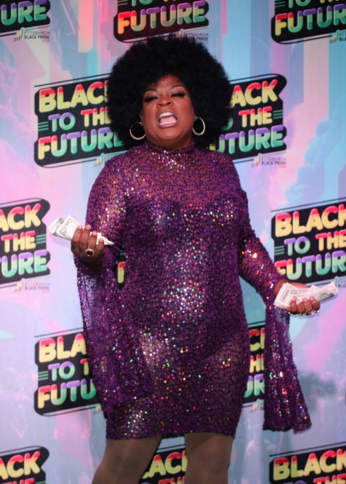 A drag queen performs in a purple dress and afro at Pittsburgh Black Pride with dollar bills in her hands.