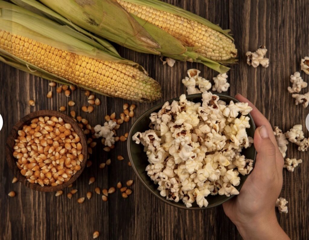 A person pushes a bowl of popcorn beside two cobs of corn and a bowl of kernels.