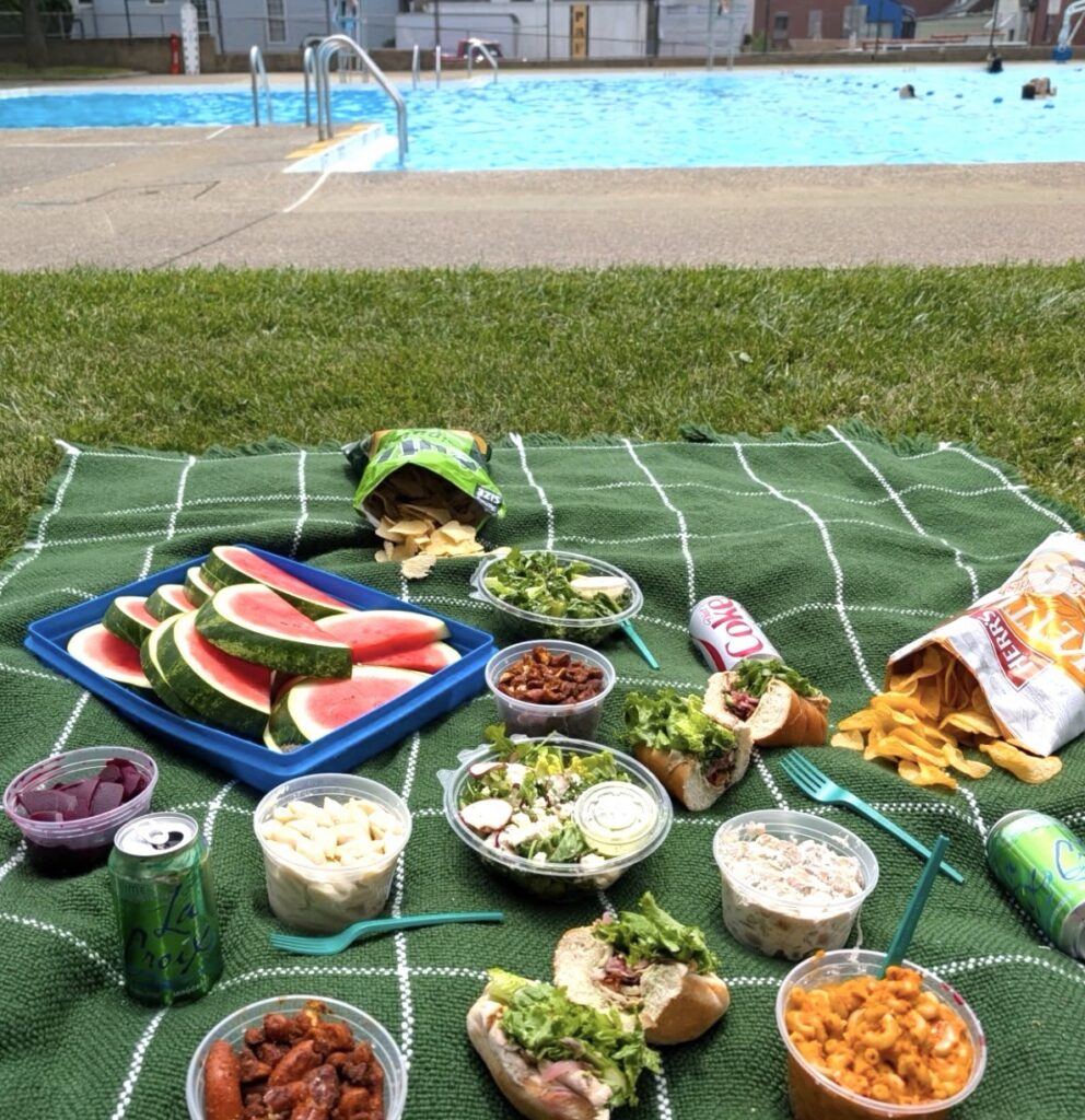 A green blanket by a pool with a picnic of salad, watermelon, and chips sitting on the blanket.