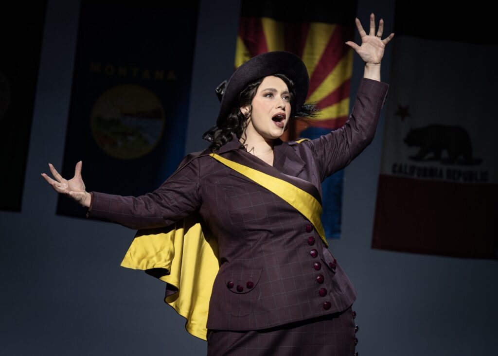 A woman in a suit and hat and yellow sash on a theater stage.