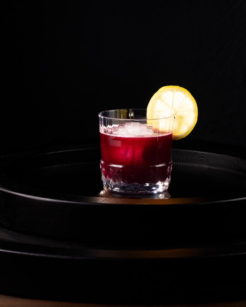A short cocktail glass with a spicy blueberry drink inside and a lemon wheel as a garnish.