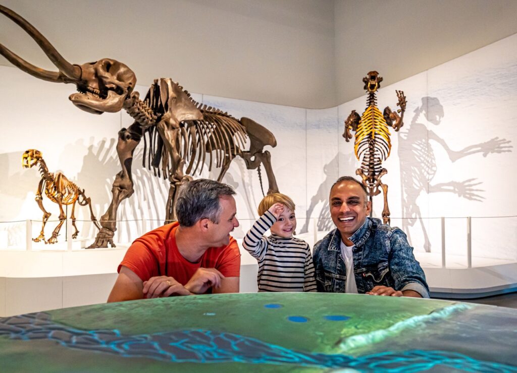 Skeleton and fossil models stand behind a family of two dads and a son.