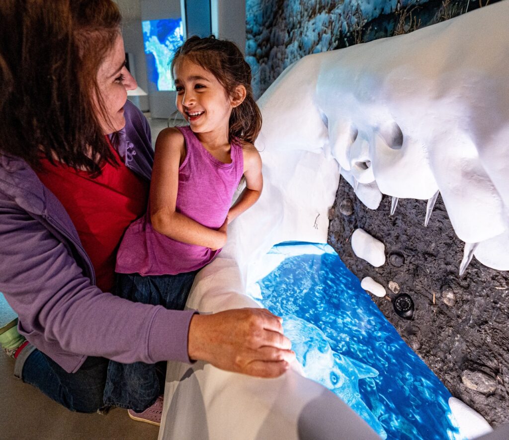 A daughter and mother look into an ice fishing hole exhibit in the Heinz History Center.