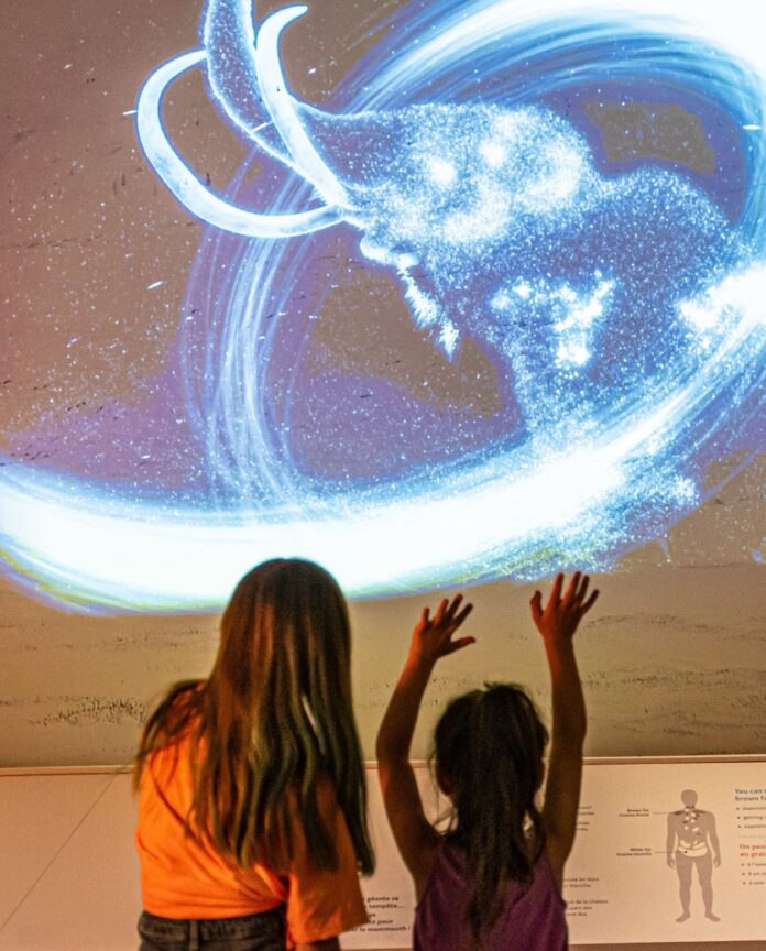 Two girls look up at a projection of an ice age mammoth on a museum wall.