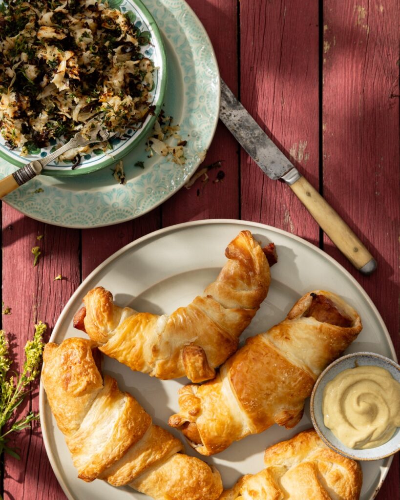 Pigs in a blanket beside a side of mustard and a bowl of sauerkraut.