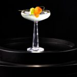 A tall coupe glass with a nearly clear spicy floral cocktail featuring a habanero and cucumber garnish.