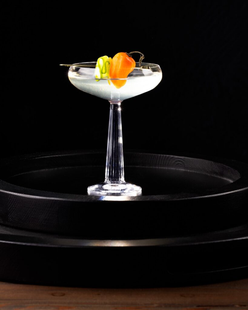 A tall coupe glass with a nearly clear spicy floral cocktail featuring a habanero and cucumber garnish.
