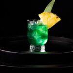 A green alcoholic punch in a short cocktail glass with ice, a pineapple slice garnish, and a pineapple leaf garnish.