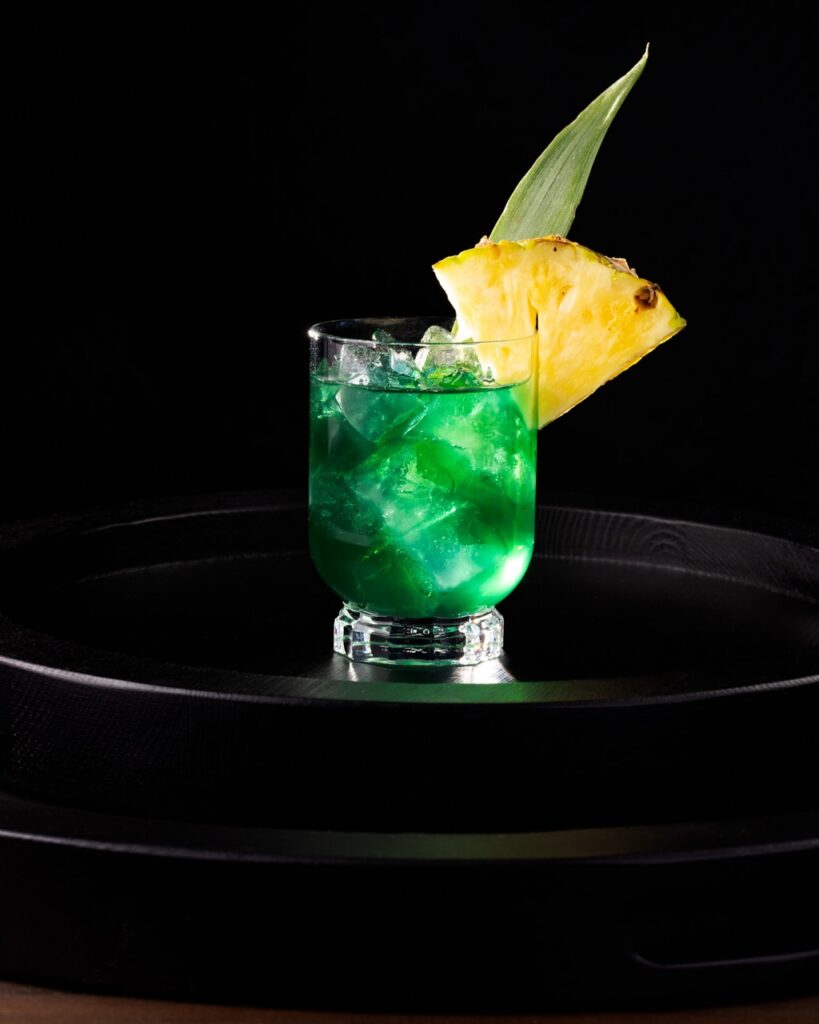 A green alcoholic punch in a short cocktail glass with ice, a pineapple slice garnish, and a pineapple leaf garnish.