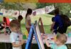 The Roving Art Cart Is One of Pittsburgh’s Best Summer Activities Two boys paint on either side of an easel with aprons on and a table of supplies.