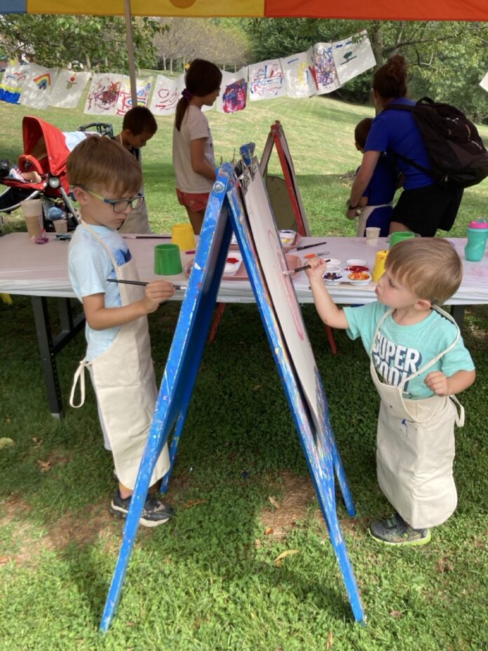 Two boys paint on either side of an easel with aprons on and a table of supplies.