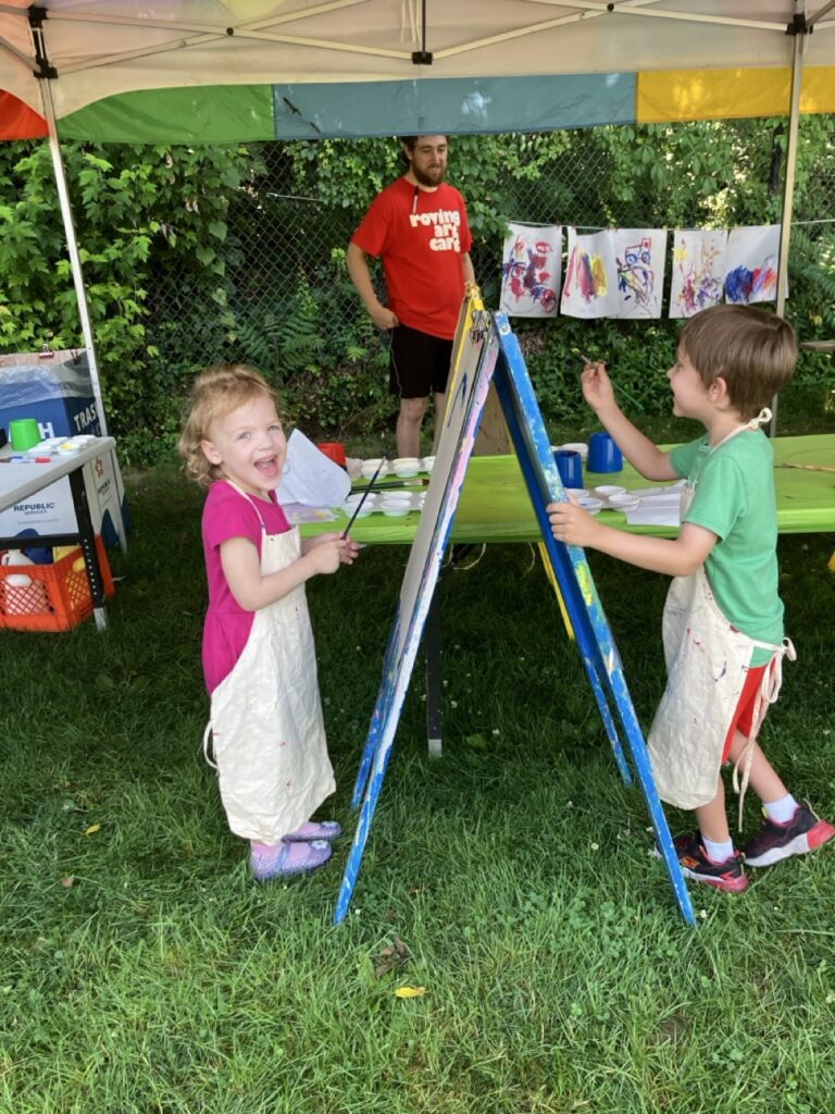 A little girl in an apron paints on an easel across from a little boy painting in an apron.