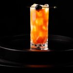 A tall glass with a chocolate-cherry gin cocktail that's orange and has a cordial cherry on a pick as a garnish.