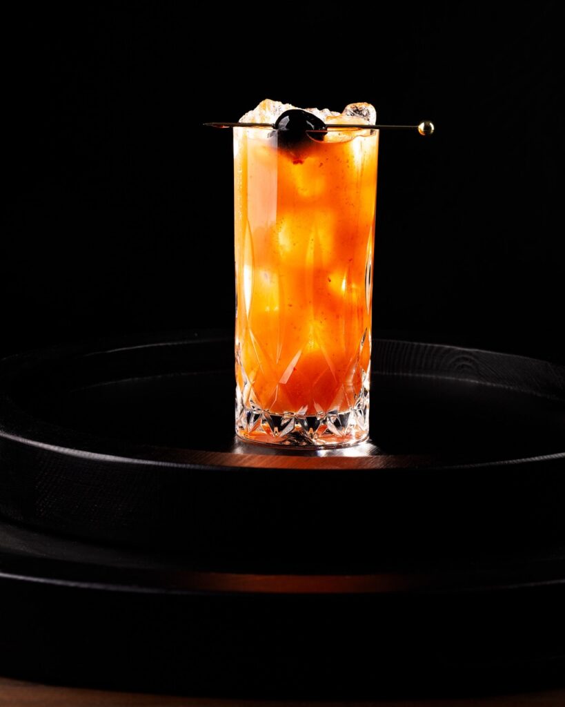 A tall glass with a chocolate-cherry gin cocktail that's orange and has a cordial cherry on a pick as a garnish.