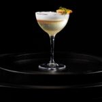 A clear clarified buttermilk punch cocktail with a white foam on top.