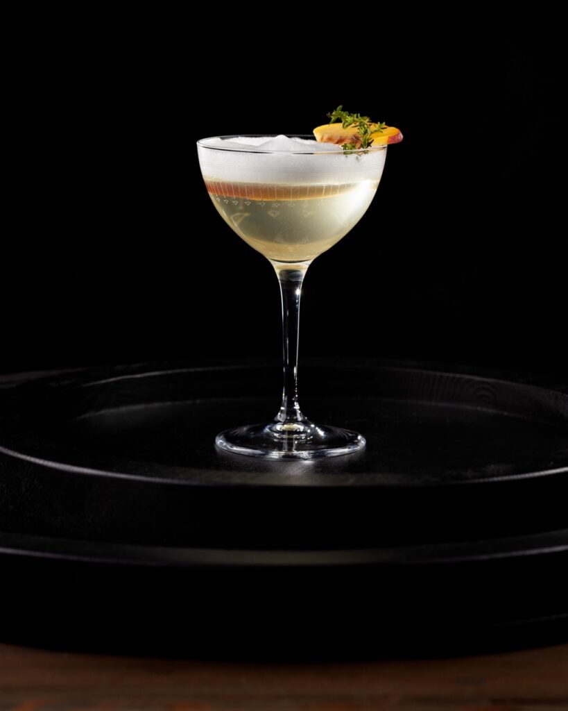 A clear clarified buttermilk punch cocktail with a white foam on top.