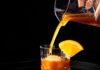 Banana Bread at Work Cocktail A banana bread cocktail pours into a rock glass with a large ice cube and orange slice garnish.