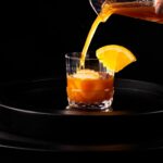A banana bread cocktail pours into a rock glass with a large ice cube and orange slice garnish.