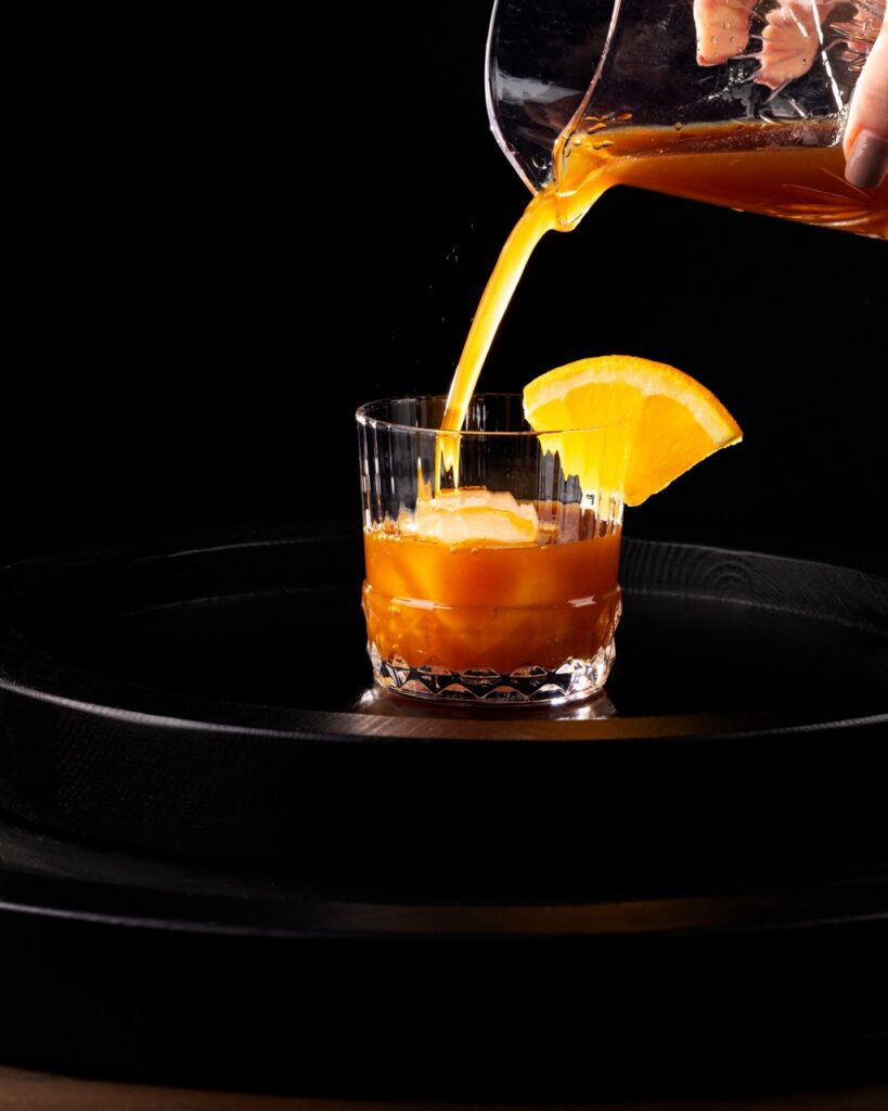 A banana bread cocktail pours into a rock glass with a large ice cube and orange slice garnish.