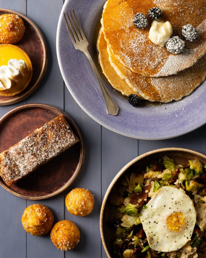 Stacks of pancakes, an egg bowl, and a slice of bread sit on a blue picnic table.