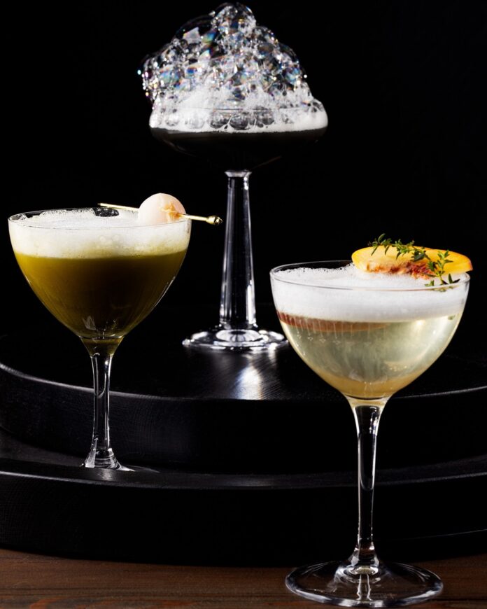 A black cocktail with foam on a top podium, a green cocktail on the second podium, and a clear cocktail with foam on the bottom podium.
