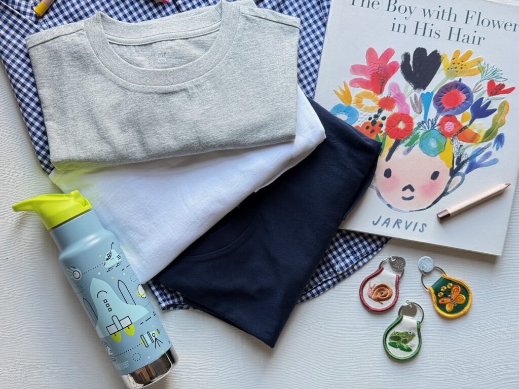 Three t-shirts in beige, white, and dark blue next to a water bottle, bag clips, and a children's book.