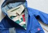 Back-to-School Shopping in Pittsburgh 2026: A Stylist’s Guide to Local Stores A dark blue rain jacket with a striped collar shirt underneath and a dark blue dinosaur backpack on top of a pair of blue jeans.