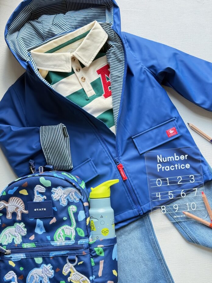 Back-to-School Shopping in Pittsburgh 2026 TABLE Magazine 3 A dark blue rain jacket with a striped collar shirt underneath and a dark blue dinosaur backpack on top of a pair of blue jeans.