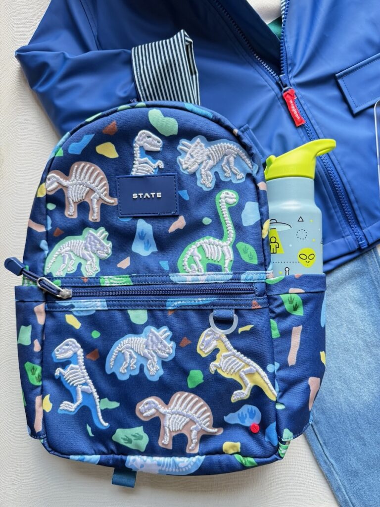 A dark blue rain jacket behind a dark blue backpack with dinosaurs and a water bottle in the pocket.